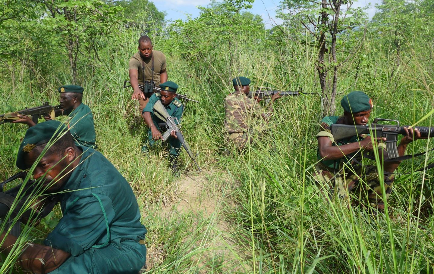 A photograph of 5 men in combat uniform with automatic rifles crouching in tall grass. A sixth man is dressed more informally and explaining something to one of the men.