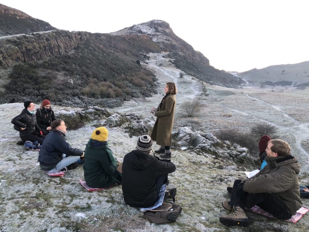 A photograph of a group of people sitting on a frosty hillside. One person is standing up and talking to the others.